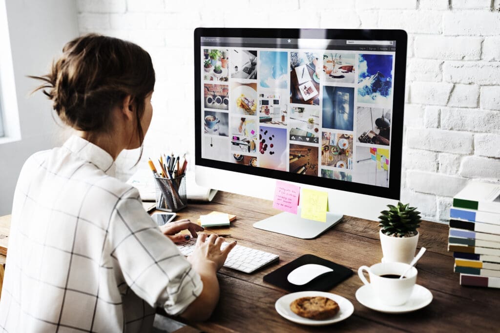 A designer sits at a wooden desk reviewing a mood board on a computer monitor in a bright, minimalist workspace.