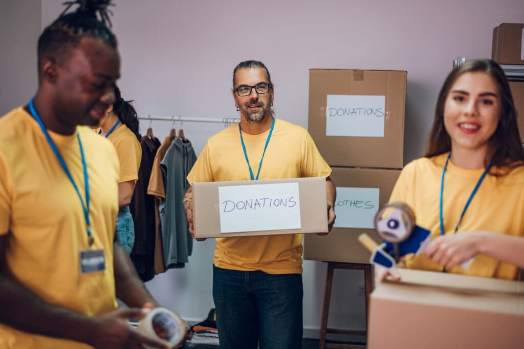 Three volunteers in yellow vests and badges sort and organize donated items and cardboard boxes in a warehouse or donation center.