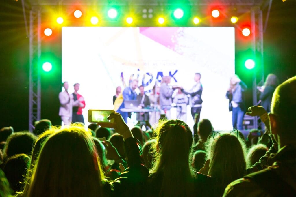 A large crowd watches performers on an illuminated stage at night with vibrant green and colorful stage lighting creating an energetic concert atmosphere.