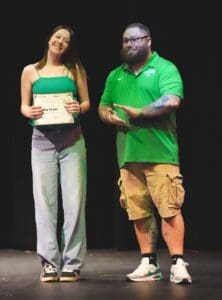 Two people stand on stage during what appears to be an awards ceremony or pageant event, with one person holding a certificate.