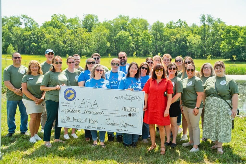 A large group of community members holds an oversized check for a charitable donation while posing together outdoors in a grassy field.