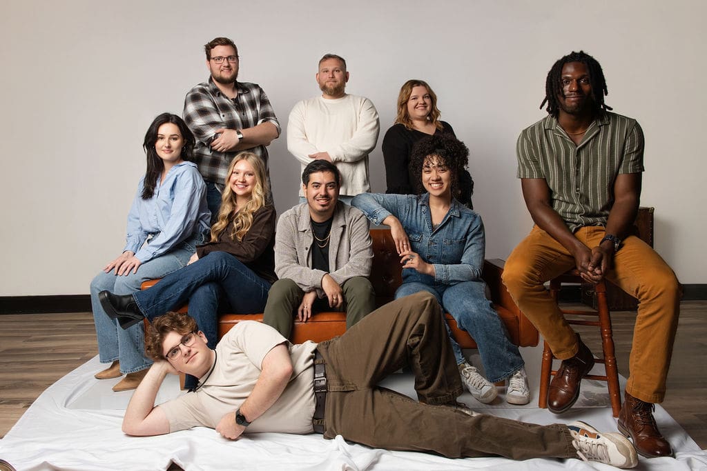 A diverse group of eight people pose together casually on a studio floor in relaxed, confident positions for a professional group portrait.